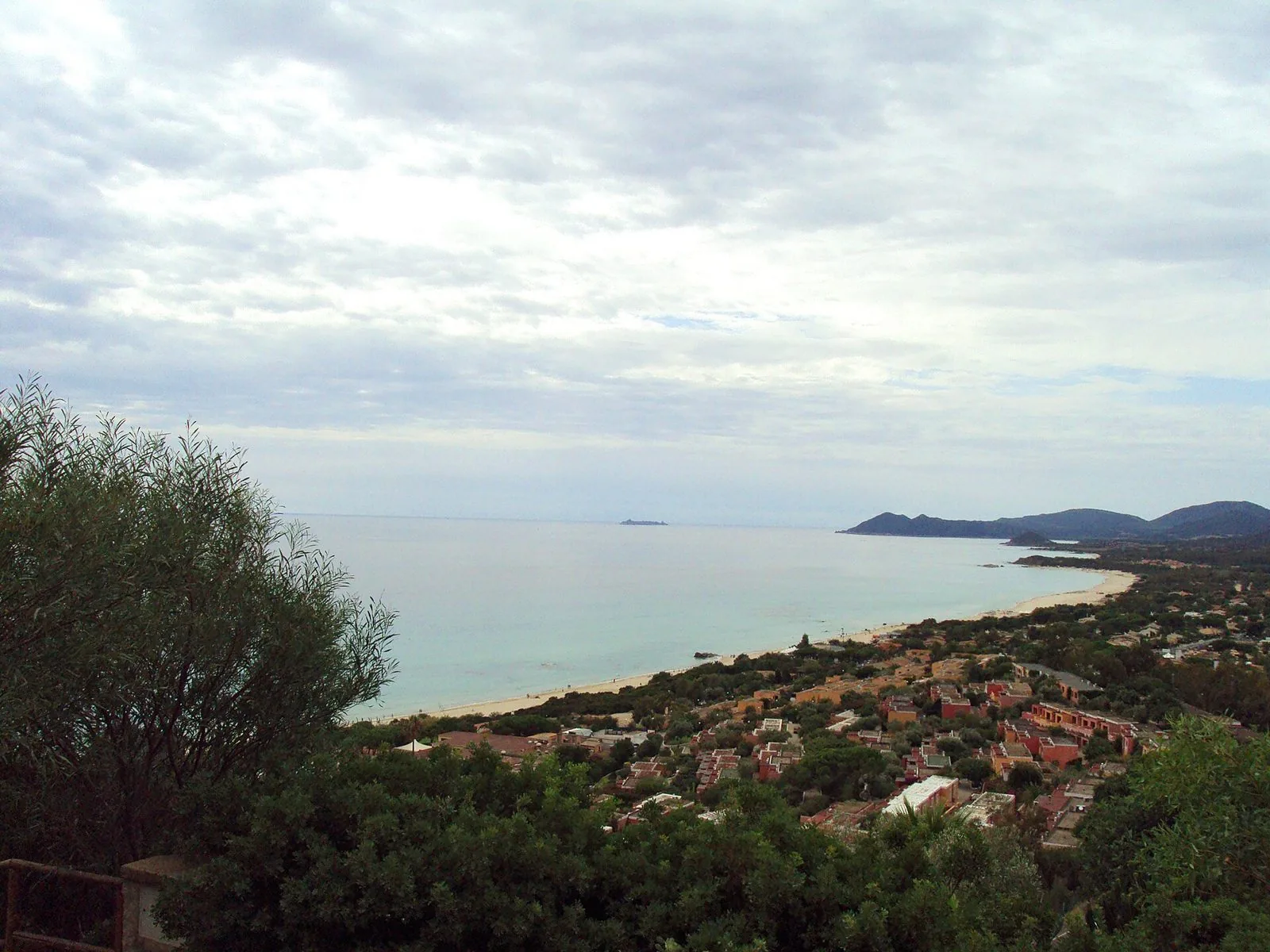 Veduta dall'alto di una lunga spiaggia chiara che curva lungo una baia dall'acqua turchese, con case tra la macchia e colline sullo sfondo